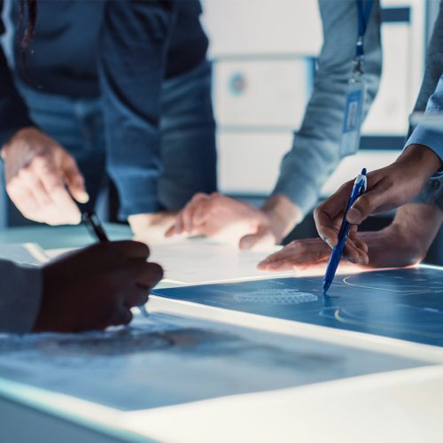 Close up of hands all collectively drawing and pointing out features on some engineering plans on a light table.